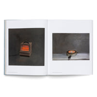 An open book on a white surface displays a two-page spread with images of vintage appliances. On the left, a brown electric space heater with glowing orange coils sits against a dark background. On the right, a closed, round, silver-colored pizzelle maker rests on a light gray surface against a dark wall.