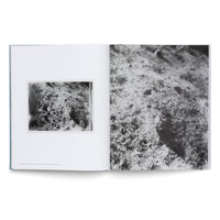 An overhead shot of an open art book on a white surface. The left page displays a small, mounted black-and-white photograph of a rough, rocky texture. The right page features a full-bleed, grainy, magnified version of the same abstract, cratered surface.
