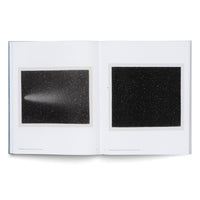 An open book on a white surface displays two black and white images of the night sky. The left page shows a comet with a long, bright tail streaking through a field of stars. The right page shows a dense, star-filled sky.