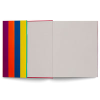 A top-down view of an open book on a white background. On the left, the fore-edge of the pages shows vibrant vertical stripes in purple, orange, blue, yellow, and green. On the right, the book is open to a blank, light gray inside cover and flyleaf, with a thin red spine visible in the crease.