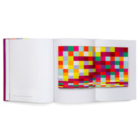An open book with a magenta spine, displayed at an angle on a white surface. The left page is primarily blank, while the right page features a colorful fold-out displaying an abstract artwork composed of a grid of vibrant squares and rectangles.