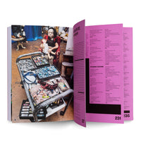 An open art book is displayed at an angle. The left page features a full-color photograph of a large, seated doll holding a smaller doll, with a metal cart full of colorful pastels and paints in the foreground. The right page contains columns of black text on a white background with a wide, bright magenta border, showing page number 231.
