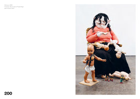A sculpture by Paula Rego, Gluttony, 2019, showing a large, seated figure wearing a grotesque, pale mask with large ears and black hair. The figure is dressed in an orange shirt and a long black skirt, with several disheveled baby dolls strewn across its lap. In front of the figure, a toddler doll stands on a platform holding a small head, while another doll lies on the floor next to miniature figurines.