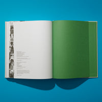 An overhead view of an open book on a bright blue surface. The left page contains publishing information next to a vertical strip of black-and-white photos. The right page is a solid, textured green endpaper, with more green pages visible behind it.