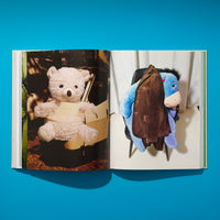 A top-down photograph of an open book on a vibrant blue surface. The left page shows a fluffy, light-colored teddy bear sitting in a dark car, held by a paper band. The right page shows a plush blue Eeyore toy on a chair against a white wall, with a brown jacket draped over it.