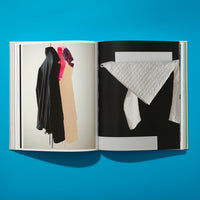 An overhead shot of an open book on a bright blue surface. The left page shows a photo of a black hooded garment and other colorful clothes hanging against a white wall. The right page has a graphic image of a white quilted fabric draped over a white bar against a black background.