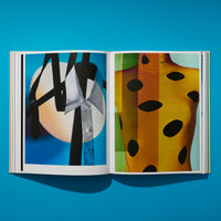 An overhead shot of an open fashion book on a bright blue surface. The left page shows an artistic photo of a white shirt collar and black ties against a glowing circle. The right page shows a mannequin in a yellow and orange striped Prada sweater with large black oval cutouts.