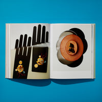 An overhead view of an open art book on a vibrant blue surface. The left page shows two ornate gold seashell brooches with pearls and gems, set against a black graphic of a stylized, blurred hand. The right page displays a circular brown leather case with a dark cameo, resting on a stack of vinyl records.