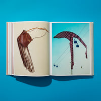 An overhead shot of an open book on a bright blue surface. The left page shows a brown leather fringed bag against a plain background. The right page shows a red plaid garment draped over a cable car line, with several gondolas visible against a clear blue sky.