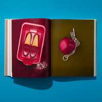An overhead shot of an open book on a bright blue surface, casting a shadow. The left page shows a pair of glossy red loafers in a red plastic bin with a matching handbag on a dark red background. The right page displays a shiny red apple with a silver chain bracelet draped over it against an olive-green background.
