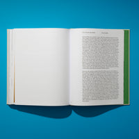 A high-angle shot of an open book with pages full of text, resting on a vibrant, solid blue background. The book is positioned slightly to the left and casts a dark shadow onto the surface below, creating a minimalist and graphic composition.