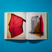 A flat lay, overhead photograph of an open book on a bright blue surface, casting a shadow. The left page shows a sheer red scarf with a black Chanel logo pattern against a yellow background. The right page features a photo of a mixed-media collage with silver chainmail, gold sequins, and red fabric.