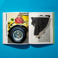 An overhead shot of an open book lying flat on a bright blue surface. The left page displays a photograph of a red handbag sitting on the back of a yellow car, with a large tire in the foreground. The right page shows a black and white polka dot garment hanging against a background of sheet music.