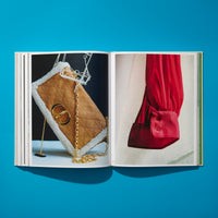 An open book lies flat on a vibrant blue surface, displaying a two-page spread of fashion photography. On the left page, a tan quilted handbag with white shearling trim and a gold chain strap is propped up. The right page shows a close-up of a silky, bright red sleeve with a buttoned cuff.