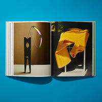 An overhead shot of an open art book lying on a vibrant blue surface. The left page displays a photo of iridescent sunglasses resting on a large, black, clothespin-like sculpture. The right page shows a photo of a bright yellow, pleated garment draped over a white frame in dramatic sunlight.