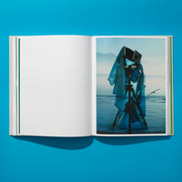 An overhead shot of an open book lying on a bright blue surface. The left page is blank, while the right page features a photograph of a large-format camera on a tripod standing in the water at the edge of a lake, with a sheer blue cloth draped over it and hazy mountains in the background.