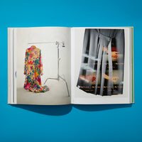 An overhead view of an open art book on a bright blue surface. The left page shows a colorful floral dress on a clothing rack against a white background. The right page features a close-up of a grey pleated skirt with an overlay of film negatives showing hands.