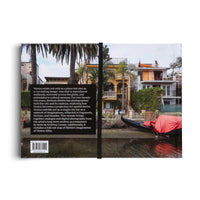 The back cover of a book featuring a photograph of a waterfront scene. A gondola with a red cover is moored in a canal in front of several houses and palm trees. One house is an ornate yellow villa, while another next to it is under construction. A black vertical band on the left contains descriptive text and a barcode.