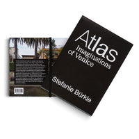 A book titled Atlas Imaginations of Venice by Stefanie Bürkle, shown on a white background. The book has a solid black cover with the title and author in a white sans-serif font. It is propped open, displaying the back cover which features a color photograph of a building with palm trees, a text description, and a barcode. A black elastic band holds the book together.