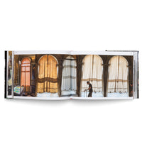 An open book laid flat against a white background, displaying a two-page color photograph of a sunlit interior. The image shows a long corridor with a series of large, arched openings covered by sheer, light-colored curtains. A man in a dark t-shirt stands in profile on the right page.