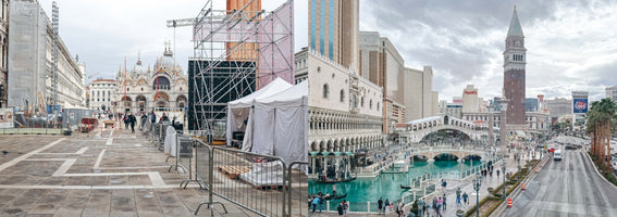 A split panoramic image contrasting the real Venice, Italy, on the left with its replica at The Venetian resort in Las Vegas on the right. The left side shows an overcast St. Mark's Square with construction scaffolding and crowd control barriers. The right side shows a bustling scene with replicas of the Doge's Palace and Rialto Bridge, a bright turquoise canal with gondolas, and the modern Las Vegas cityscape in the background.