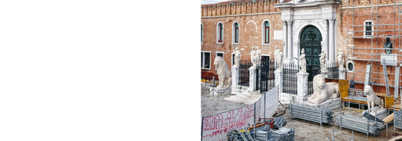 The historic main gate of the Venetian Arsenal, featuring an ornate white marble entrance on a red brick building, is undergoing restoration. The area is filled with scaffolding, temporary fencing, and stacks of construction materials, partially obscuring the large marble lion statues that flank the entrance.