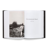 A studio shot of an open, black hardcover book on a white background. The left page features a black-and-white photo of a man in a light suit and fedora walking in a field near a horse. The right page is a chapter title in German: Der monumentale Munch (1909–1914).